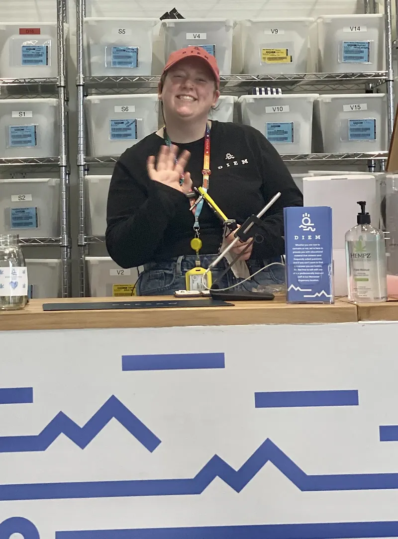 A Diem Lynn budtender smiling and waving from behind the counter